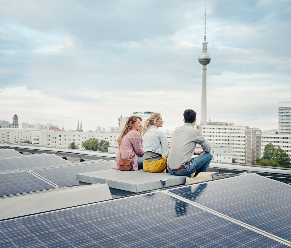 Drei Personen sitzen auf einem Dach mit einer Solaranlage mit Blick auf den Fernsehturm