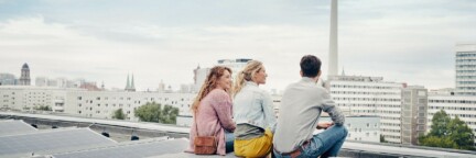 Drei Personen sitzen auf einem Dach mit einer Solaranlage mit Blick auf den Fernsehturm