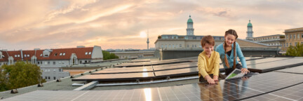 Zwei Kinder sitzen auf einem Dach mit einer Solaranlage der Berliner Stadtwerke. Im Hintergrund ist ein Sonnenuntergang und der Berliner Fernsehturm zu sehen.