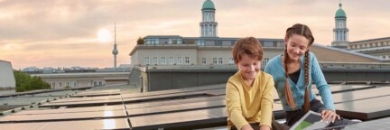 Zwei Kinder sitzen auf einem Dach mit einer Solaranlage der Berliner Stadtwerke. Im Hintergrund ist ein Sonnenuntergang und der Berliner Fernsehturm zu sehen.