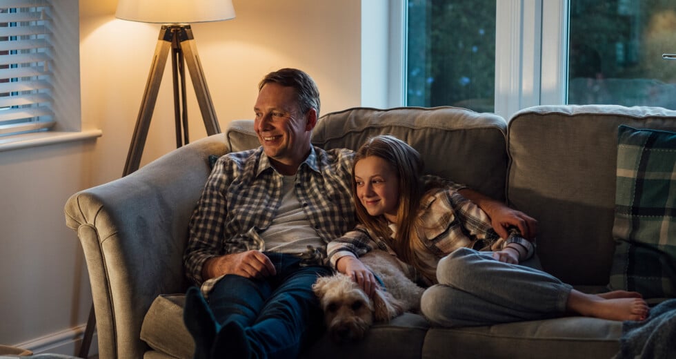 Vater sitzt mit Tochter und Hund auf dem Sofa und sieht fern