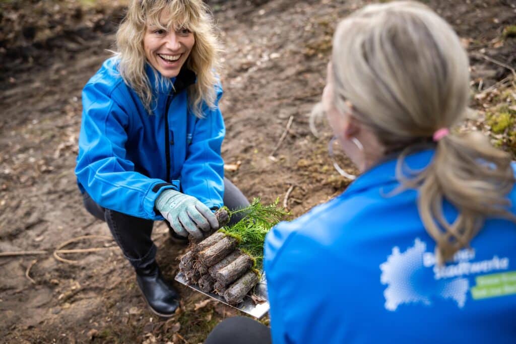Zwei lächelnde Mitarbeiterinnen der Berliner Stadtwerke knien auf dem Waldboden und halten Setzlinge auf einem Spaten für die Pflanzaktion im Stadtwerke-Wald.