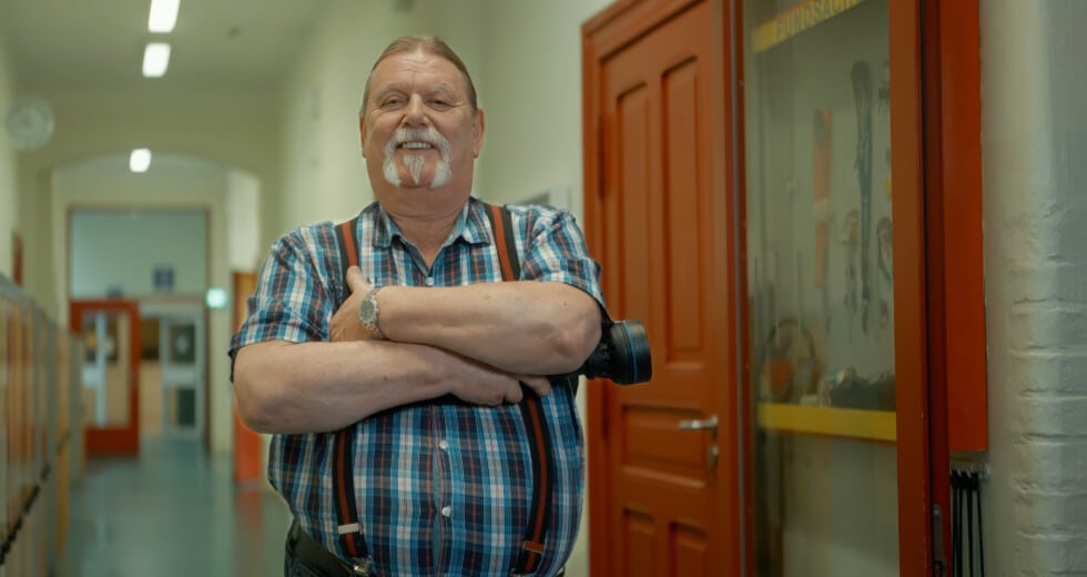Ein Hausmeister steht in einem Schulflur und blickt lächelnd in die Kamera.